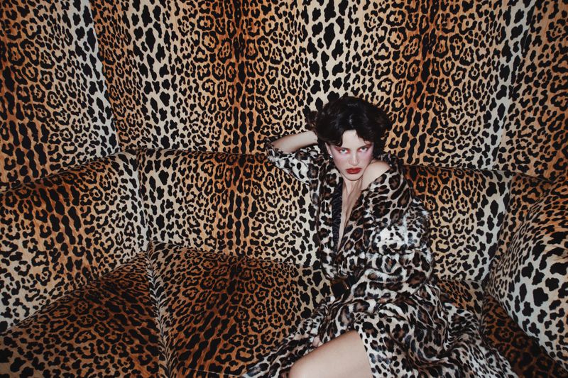 Brunette adult female in leopard-print robe poses on matching leopard-print sofa with a leopard-print wall in the background.