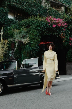 Elegant woman in yellow skirt suit and red heels walking on street next to black convertible car with ivy covered building in background.