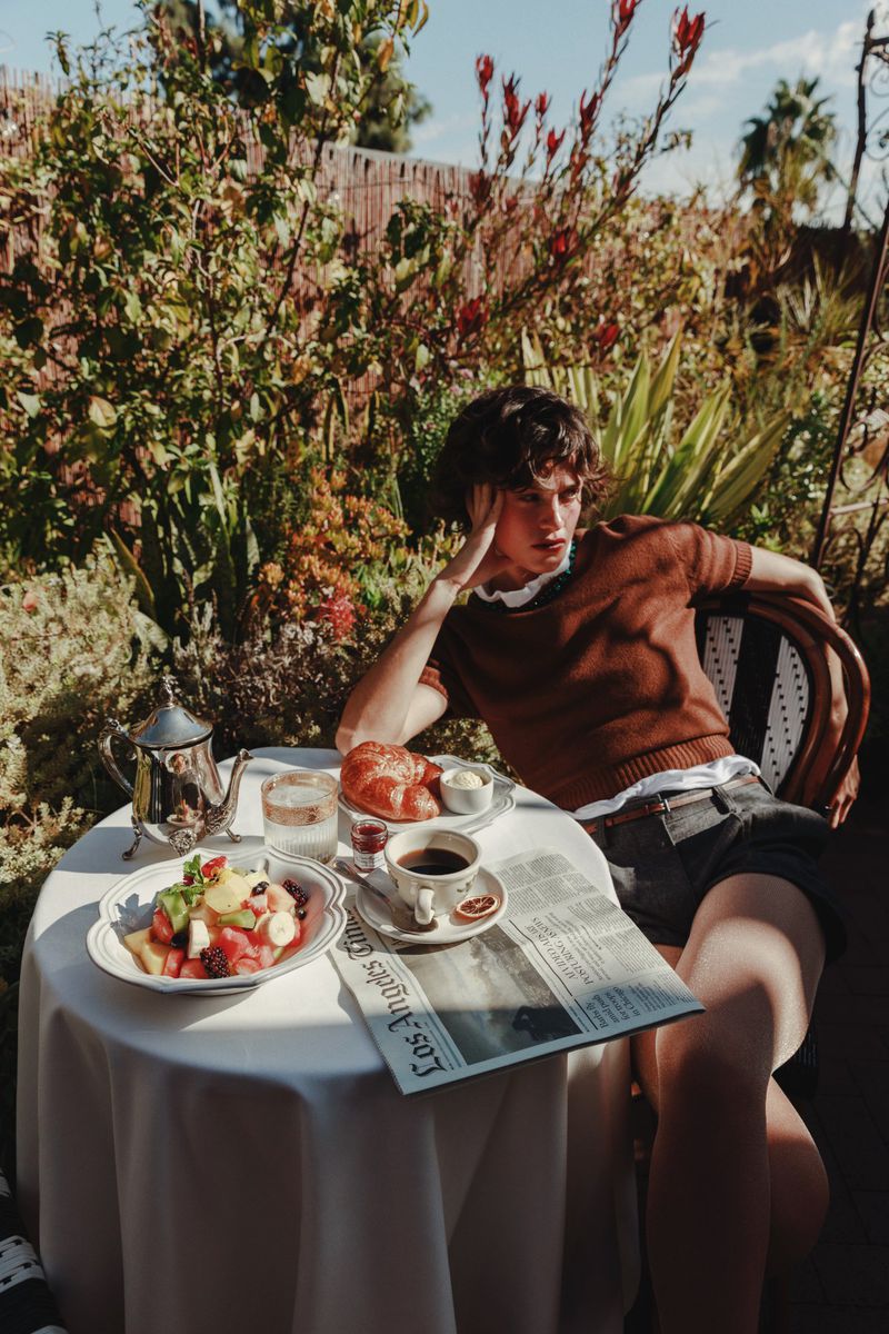 Woman sits at outdoor table with breakfast in garden setting, reading Los Angeles Times newspaper on a sunny day.