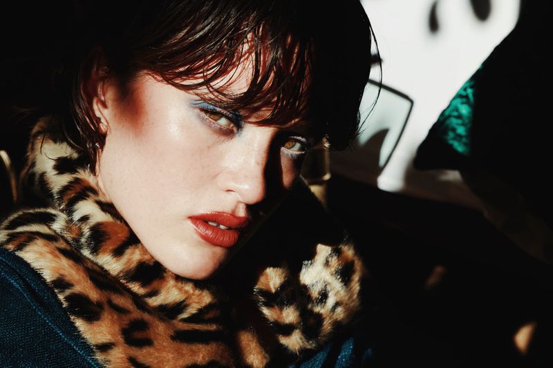 Close-up of a woman with wet dark hair, striking blue eyeshadow, and red lipstick, wearing an animal print collar in a dark setting.