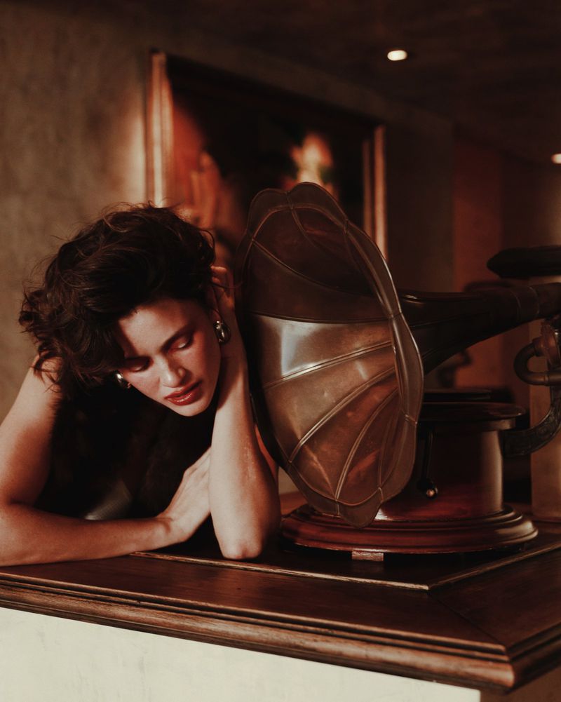 Woman with dark curly hair rests her head on her arms next to a vintage phonograph in a warm-toned interior room with painting.