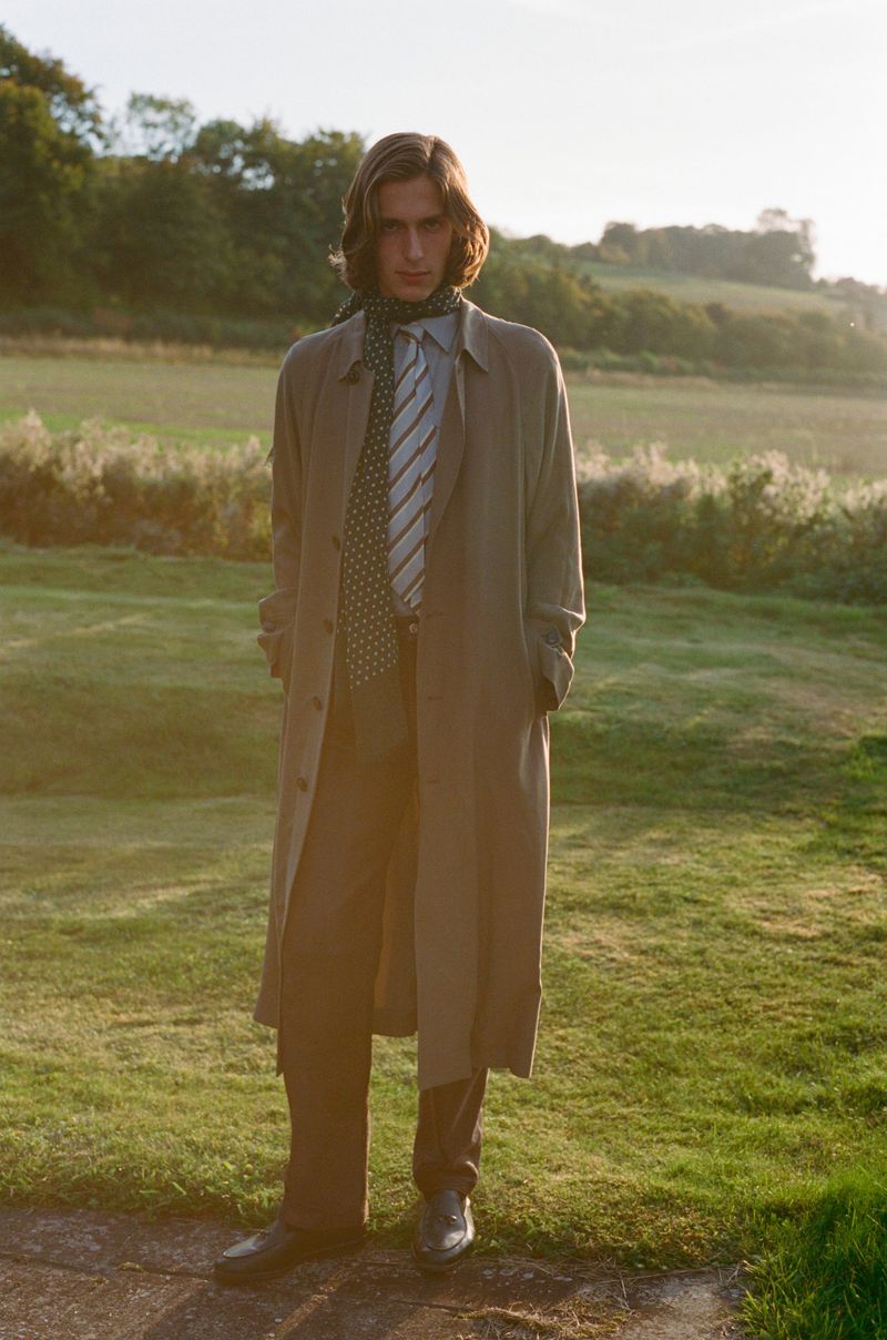 Male fashion model in trench coat stands in green field with necktie, styled hair, and black shoes in soft sunlight.