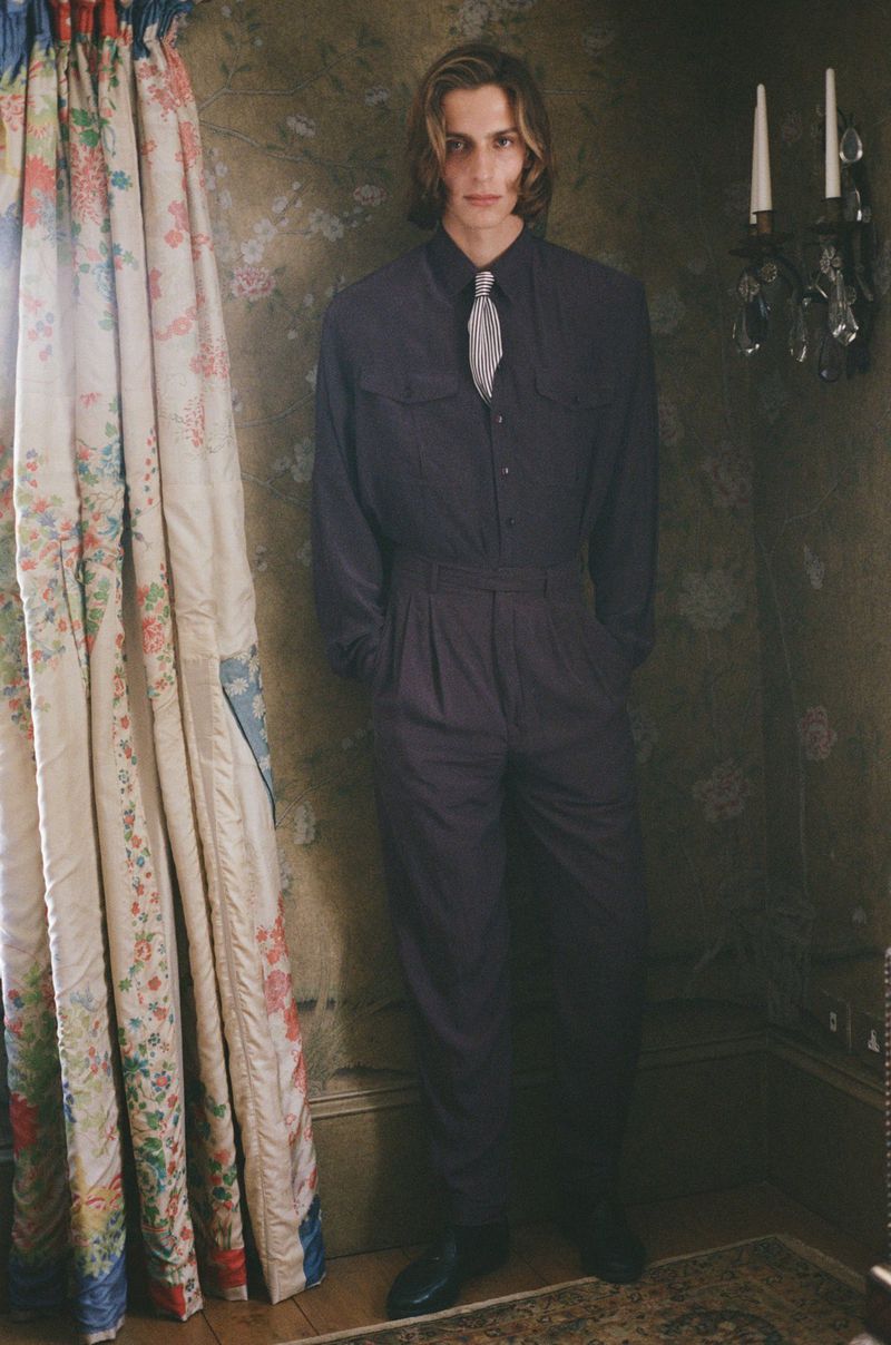 Adult male in dark blue jumpsuit and silver tie standing indoors near floral curtains and a decorative sconce on a patterned wall.
