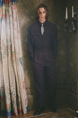 Adult male in dark blue jumpsuit and silver tie standing indoors near floral curtains and a decorative sconce on a patterned wall.