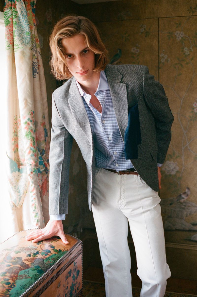 Adult male posing indoors wearing blazer, button-down shirt, and trousers leaning against antique trunk with floral curtains in the background.