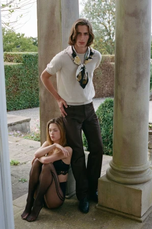 Elegant fashion photo shoot outdoors with a stylishly dressed couple posing by stone columns and manicured hedges in the background.