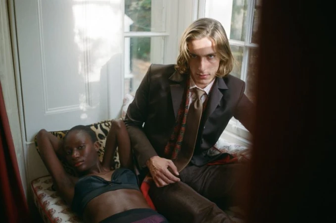 Two models pose indoors near a window: a woman reclines on a floral chaise lounge while a man sits beside her in a brown suit.