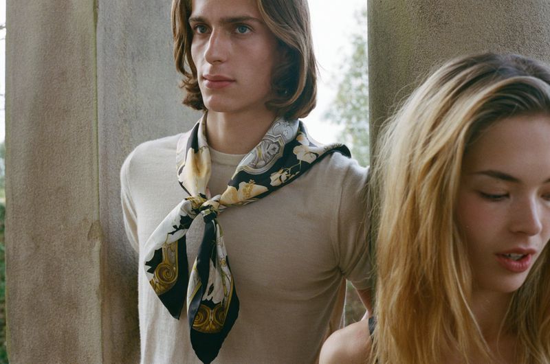 A close-up of a stylish adult couple in an outdoor setting. The man wears a patterned scarf, exuding a blend of fashion and editorial style.