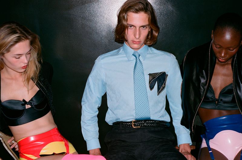 Three fashion models pose in a studio with bright latex outfits and bold accessories against a dark backdrop, highlighted by cool, moody lighting.