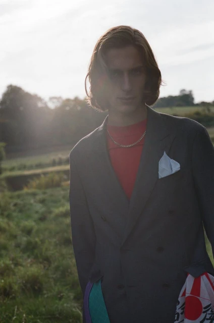 Adult male in a gray suit and red top posing in a field with sunlit sky, wearing a gold chain necklace and pocket square.