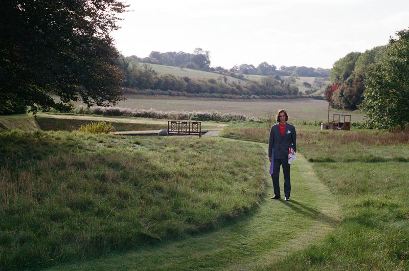Adult male in suit standing on grassy path in rural landscape with trees and bridge under bright sky in outdoor setting.