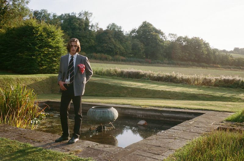 A young adult man in a gray blazer, tie, and dark pants poses by a reflective pool in a grassy outdoor setting with trees and shrubs.