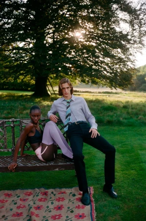 Deborah Akanni and a male model posing beside an ornate iron bench in a grassy field with a large tree in the background.