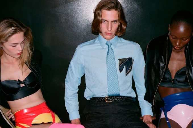 Three models seated against a dark background, the central man in a light blue shirt and tie flanked by women in latex and lingerie.