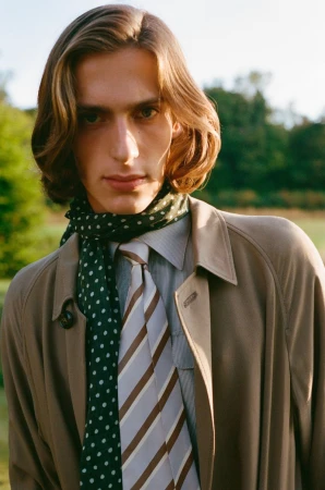 Close-up portrait of a male model with long hair wearing a beige coat, striped tie, and green polka dot scarf outdoors.