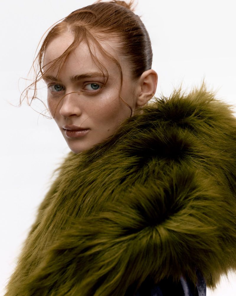 Close-up shot of a redhead woman with fair skin in a green fur coat with an expressionless face against a white background.