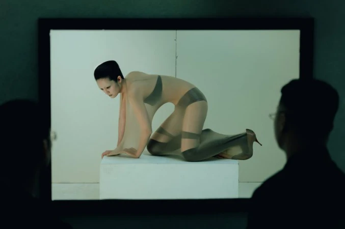 Asian woman in a sheer bodysuit posing on a white platform in front of a white background, observed by two people with dark silhouettes.