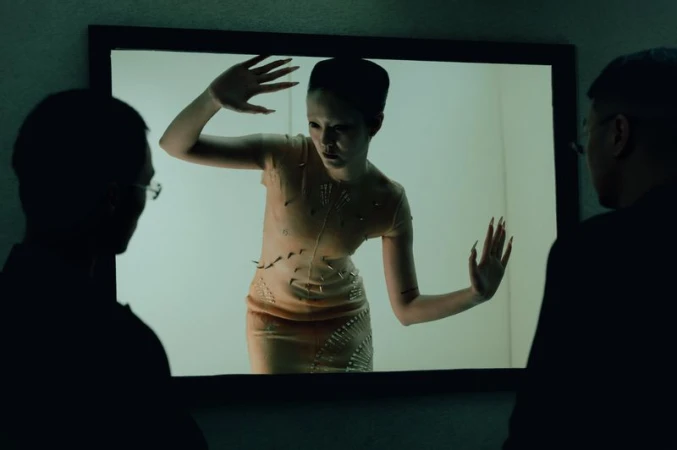 Fashion image displayed on screen. Two men in dark silhouettes view the image, which features a model in a spiked dress.