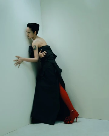 Woman in a strapless black dress and red tights leans against a corner. Studio lighting. Fashion editorial shot.