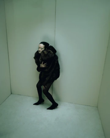 Woman in fur coat and heels stands in the corner of an empty room with green lighting and blank walls, creating a minimalist fashion shot.