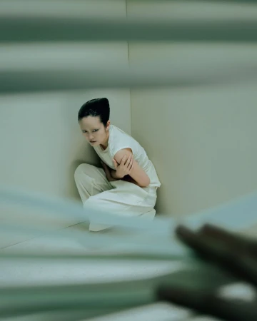 A person crouches in a corner with arms crossed, wearing white top and pants in a minimalist indoor setting, creating a mood of discomfort.
