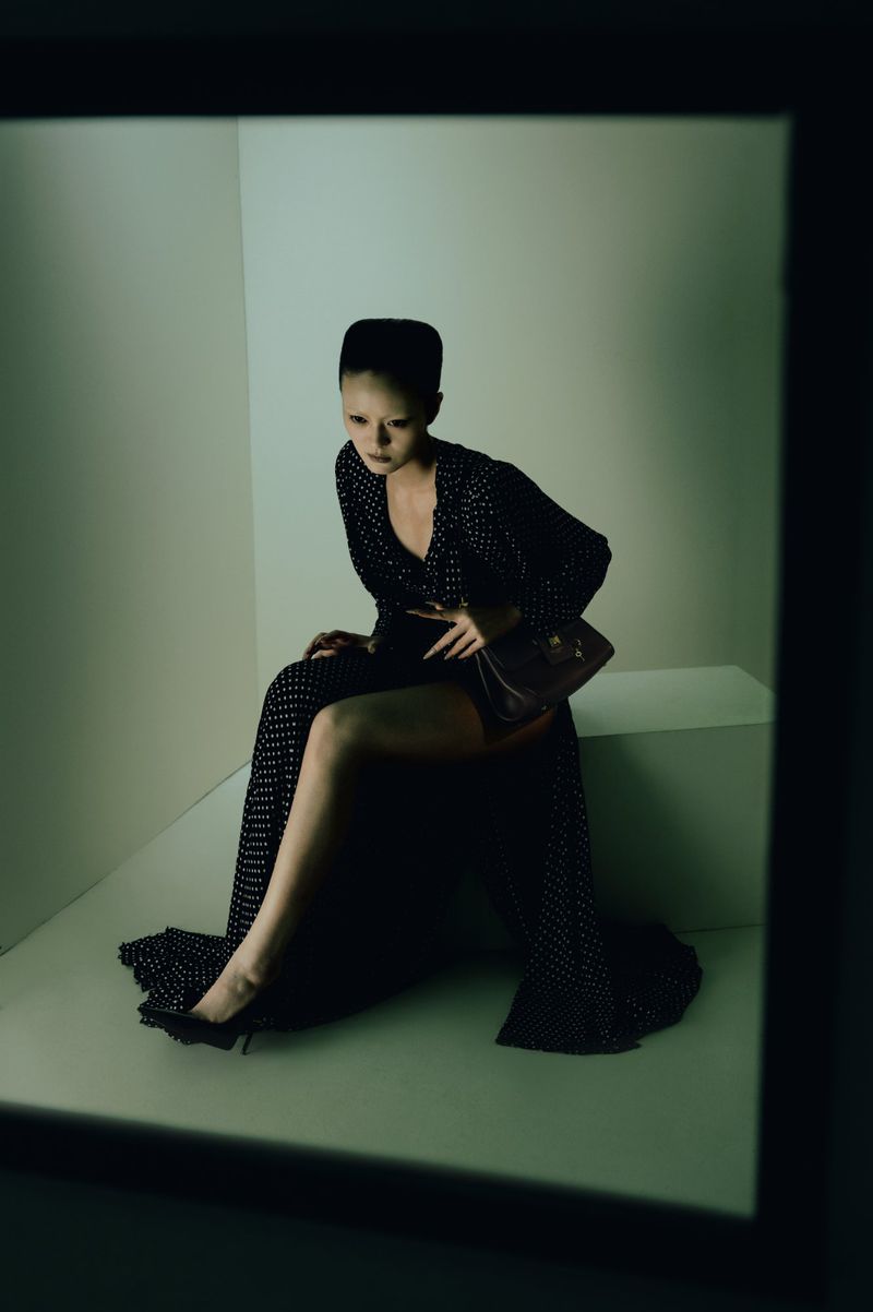 Elegant woman in a polka dot dress and heels, seated indoors with a handbag, posing in a square frame with soft, diffused lighting.