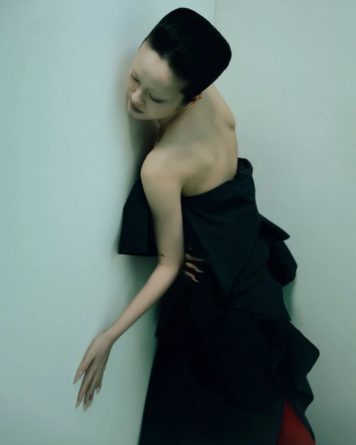 Model in a black strapless gown leaning against a light green corner wall in a studio portrait.