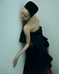 Model in a black strapless gown leaning against a light green corner wall in a studio portrait.