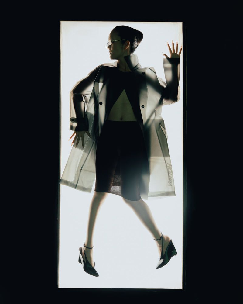 Model Yvette Chau wearing a translucent trench coat and black heels silhouetted against a glowing rectangular light panel.