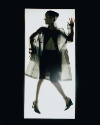 Model Yvette Chau wearing a translucent trench coat and black heels silhouetted against a glowing rectangular light panel.