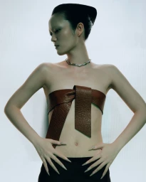 Model Yvette Chau wearing a textured brown leather bow top and silver necklace against a white illuminated background.