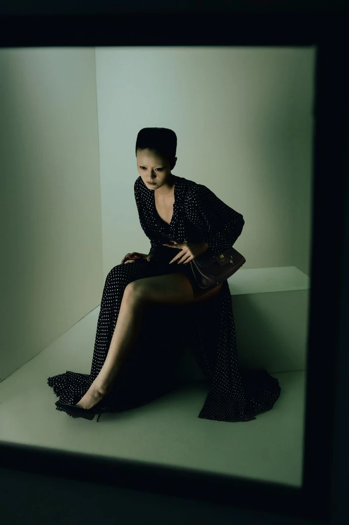 Model Yvette Chau wearing a black patterned gown and high heels, seated inside an enclosed geometric box set.