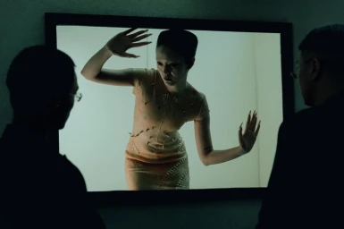 A model inside a lit frame poses with raised hands while two silhouetted figures watch from the foreground.