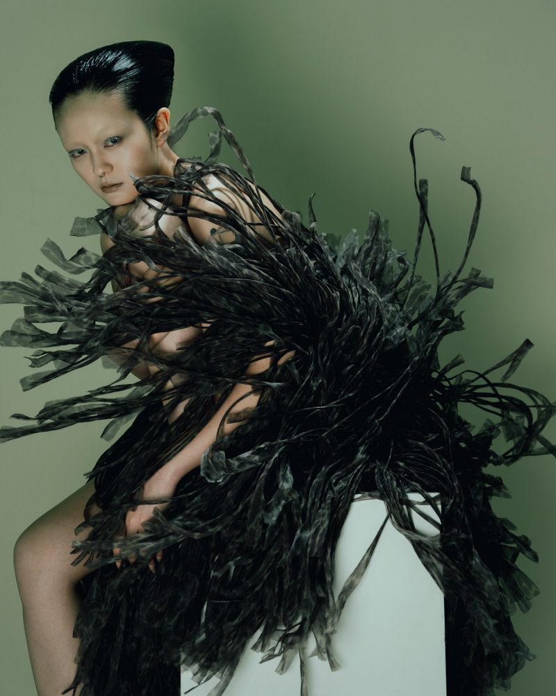 Model Yvette Chau posing in a voluminous black fringed sculptural dress against a muted green studio backdrop.