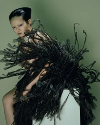 Model Yvette Chau posing in a voluminous black fringed sculptural dress against a muted green studio backdrop.