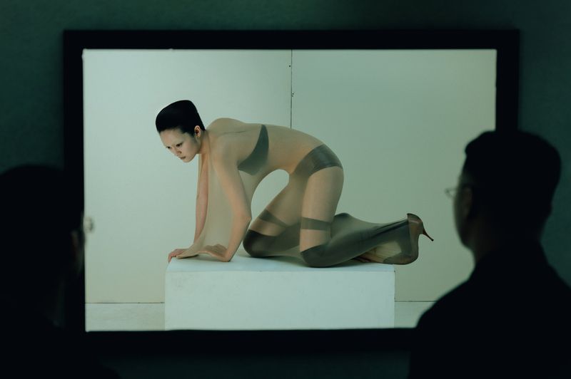 Model Yvette Chau poses on all fours on a white plinth wearing sheer beige nylon, framed on a display screen between two silhouettes.