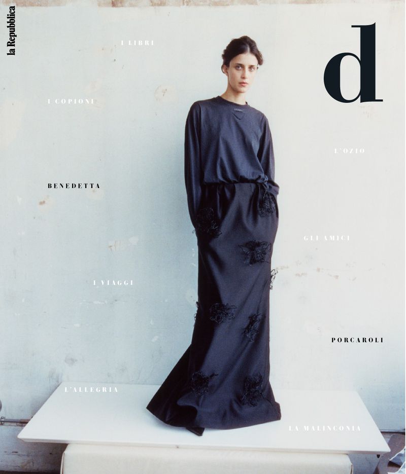 Magazine cover featuring a female model wearing a long black dress, posing on a white platform in front of a textured wall with type.