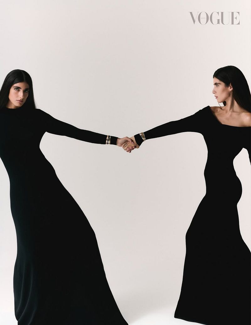 Two women in black dresses hold hands in a studio setting for a Vogue editorial, against a neutral backdrop, showcasing fashion and unity.