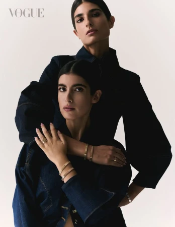 Two women in denim jackets pose intimately against a bright backdrop, jewelry and makeup accentuating their stylish, fashionable looks.