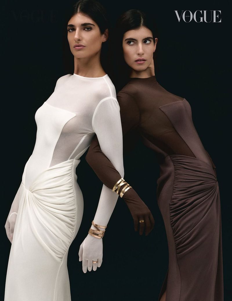 Two women in couture gowns pose against a dark backdrop, adorned with gold bangles, exuding elegance and sophistication in a fashion editorial.