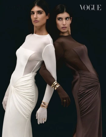 Two women in couture gowns pose against a dark backdrop, adorned with gold bangles, exuding elegance and sophistication in a fashion editorial.