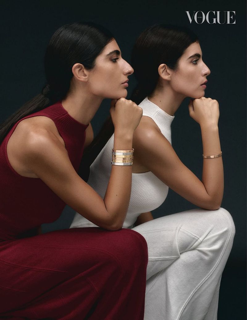 Two adult women pose in studio wearing sleeveless tops and slacks, accessorized with gold bracelets, set against a dark background.