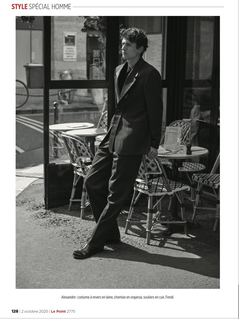 A man in a dark suit stands beside a sidewalk cafe with patterned chairs. A black and white fashion editorial image.