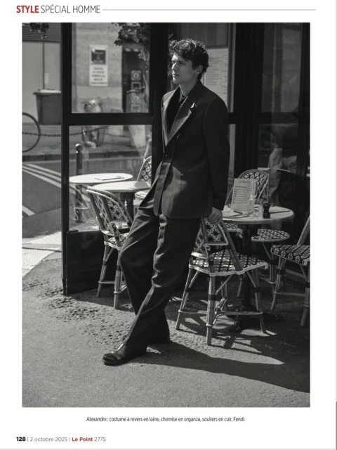 A man in a dark suit stands beside a sidewalk cafe with patterned chairs. A black and white fashion editorial image.