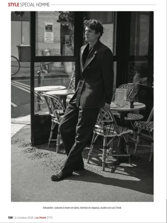 A man in a dark suit stands beside a sidewalk cafe with patterned chairs. A black and white fashion editorial image.