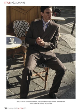 Adult male poses in a brown double-breasted suit outside on a sunny day, seated on a wicker chair at a bistro table with a coffee cup.