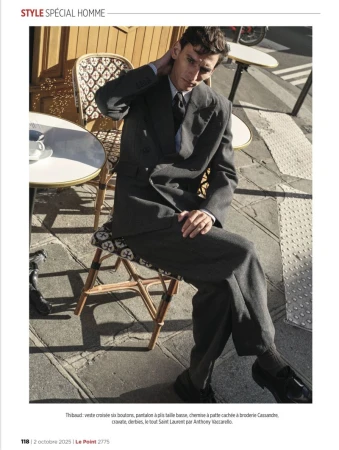 A man in a grey suit sitting on a woven chair outside a cafe, with white tables and metal chairs around him in daytime.