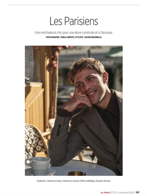 Portrait of a smiling man wearing a grey suit in an outdoor cafe. The man sits at a white table next to a cup and small dish.