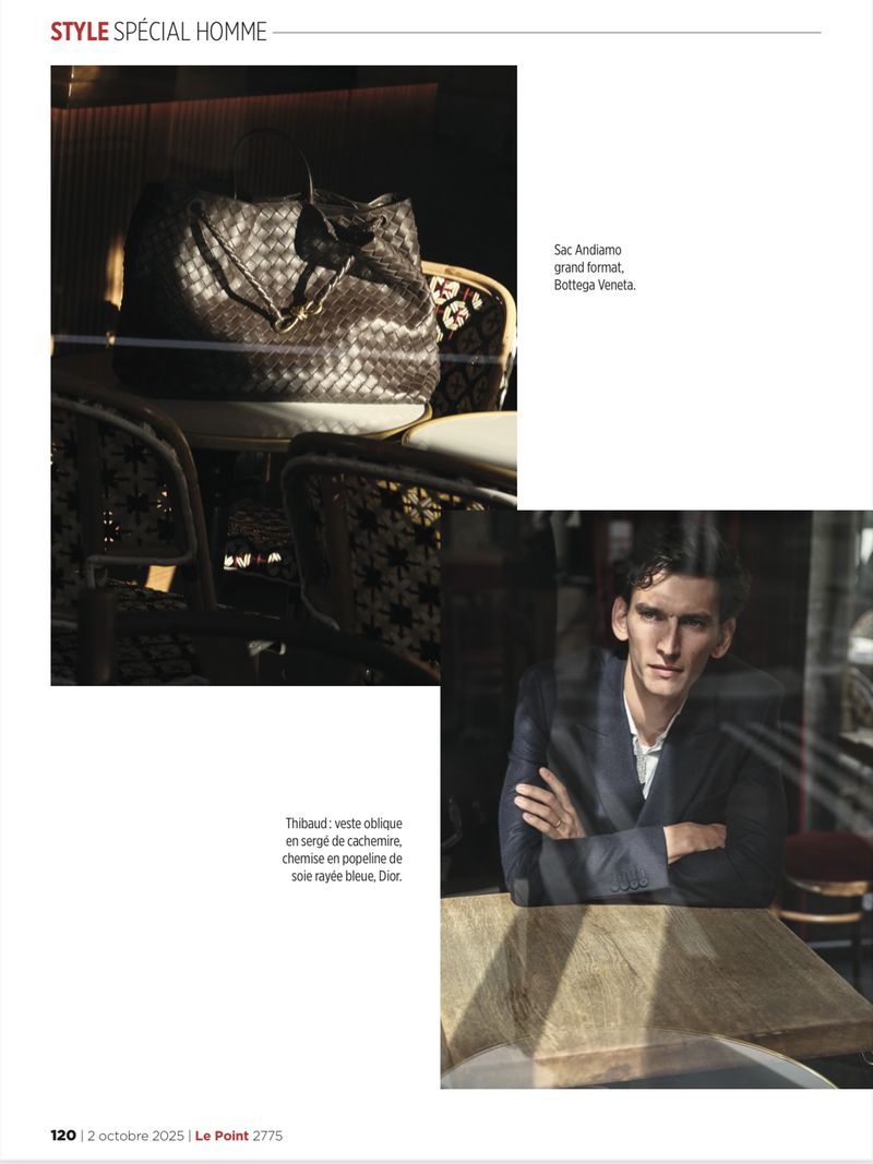 Editorial fashion shot of a stylish man in a blazer with a designer bag on a chair inside an urban cafe, illuminated by window light.