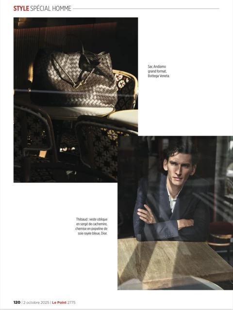 Editorial fashion shot of a stylish man in a blazer with a designer bag on a chair inside an urban cafe, illuminated by window light.